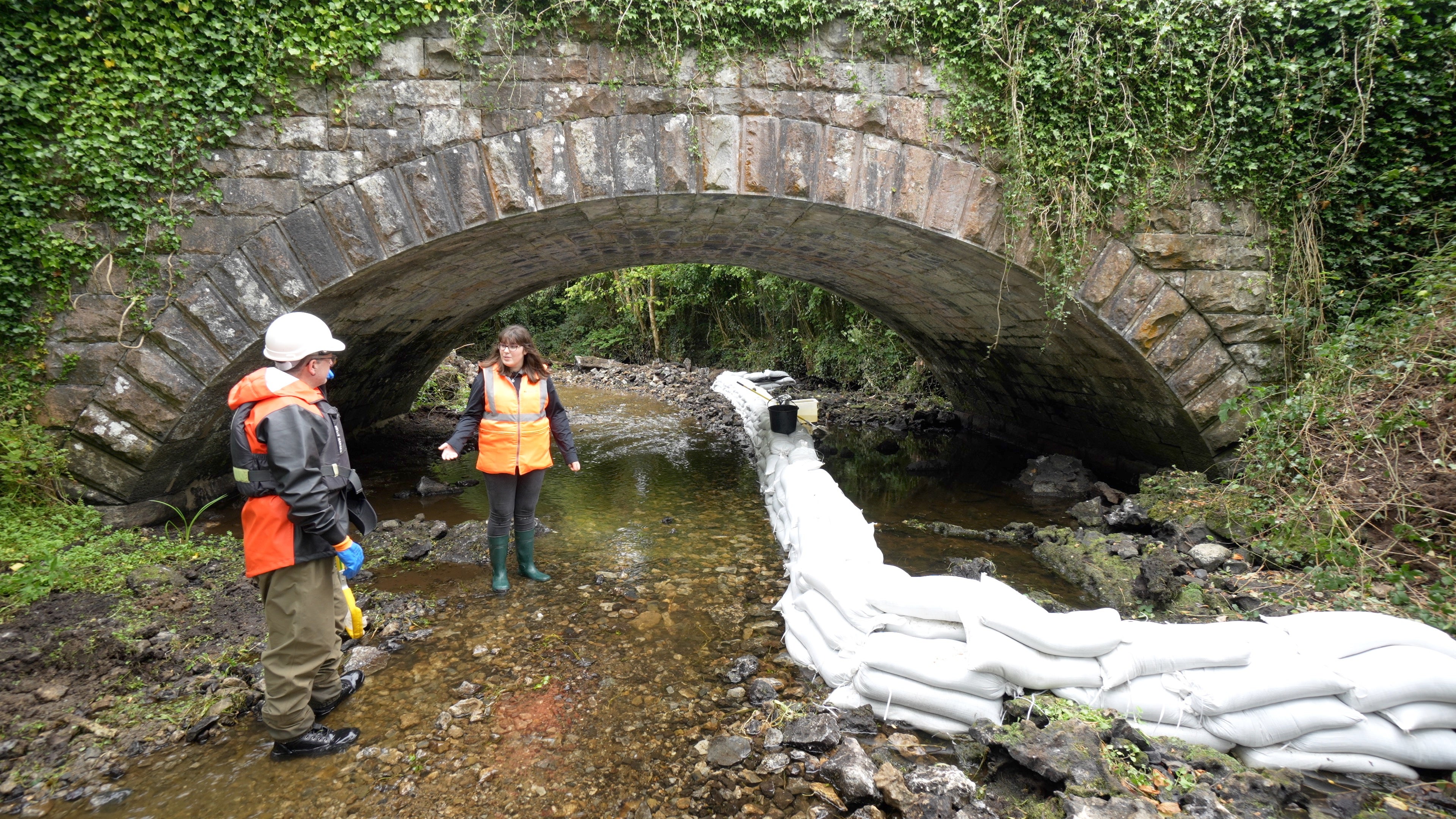 the image provided shows two Iarnród Éireann employees in high-visibility safety gear and life jackets working near a stone bridge arch in a stream.